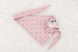 A pink pet bandana with a repeating geometric star pattern and a decorative printed corner patch, laid flat on a white textured faux fur background.